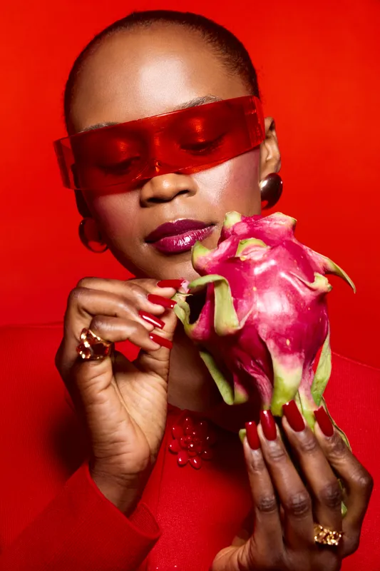 Elegant black woman with dark skin holds a dragon fruit against a red background. She wears red sunglasses and red nail polish, creating a bold fashion and beauty concept.