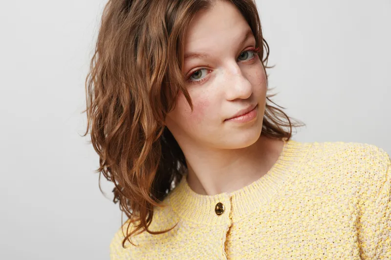 Young woman with wet brown hair wearing yellow knitted sweater looks at camera with soft expression on grey background. Concept of natural beauty and casual style.