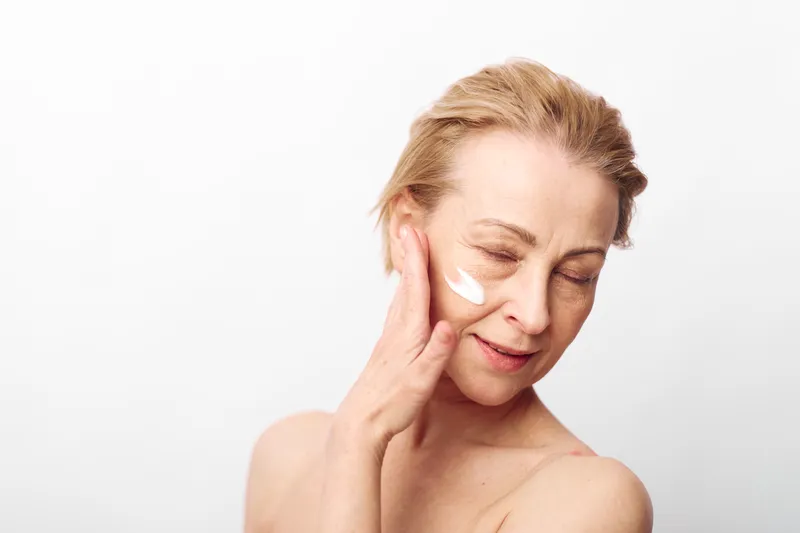 Mature woman with short blonde hair applies white face cream on cheek. Soft smile with closed eyes on plain white background. Concept for skincare, anti aging, wellness, and self care.