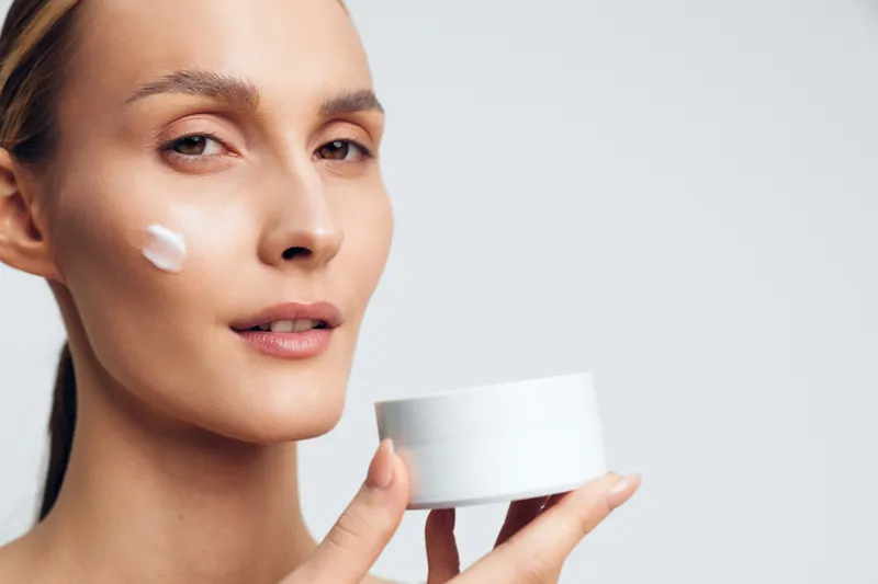 Young woman applying moisturizer on face holding white jar skincare cream closeup, natural light, smooth skin texture, beauty routine, neutral background.