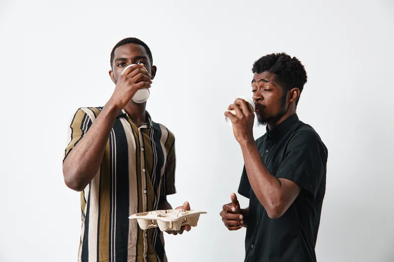 Two young black men with dark skin drinking coffee from disposable cups, casual clothing, standing together on white background, enjoying beverage and conversation.