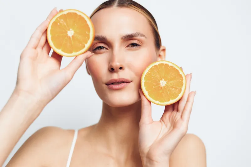 Young woman holding two halves of fresh orange fruit near her face. Close-up on white background. Concept of healthy lifestyle, skincare, natural beauty, and vitamin C.
