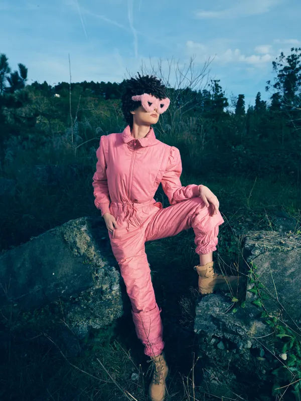 Fashion model in pink jumpsuit and mask poses on rocky terrain outdoors, dramatic look and bold stance, creative portrait for editorial or creative project in nature setting.