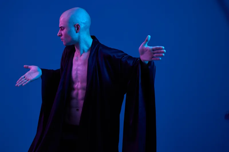 Strong man poses dramatically in a flowing black robe under dramatic blue lighting, showcasing strength and motion. Captivating and powerful portrayal of confidence.