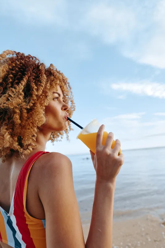 Young woman with curly hair drinks orange juice through a straw while standing on a sandy beach under a partly cloudy sky during a warm day.
