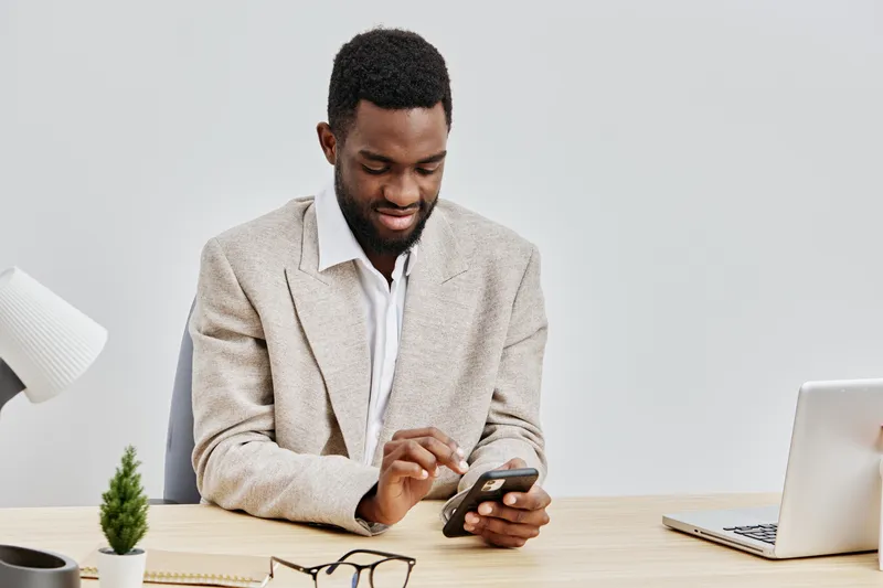 Professional young African American man sitting at desk, wearing beige blazer and white shirt, using smartphone, smiling, in modern office environment with laptop, glasses, and small plant on white ba