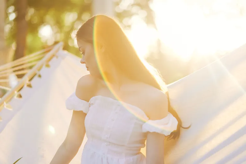 Young woman in a white dress enjoying sunlight, with a dreamy expression, surrounded by greenery and soft light reflections in spring season
