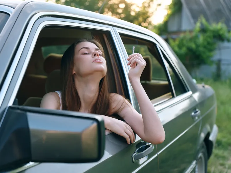Young woman enjoying a moment of relaxation with a cigarette while sitting in a car, surrounded by nature, evoking a sense of freedom and introspection.