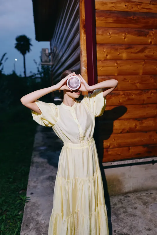 Portrait of a woman in a pale yellow dress posing outdoors beside a wooden building at dusk, covering her face with a round camera for a playful moody street style.