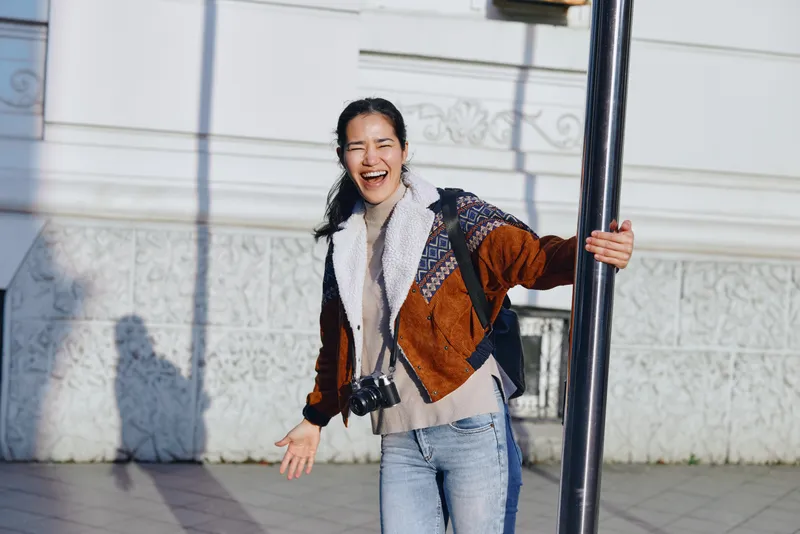 Joyful woman outdoors, laughing beside a street pole with a camera hanging from her neck, casual jacket and scarf, candid moment of happiness and exploration in urban daylight