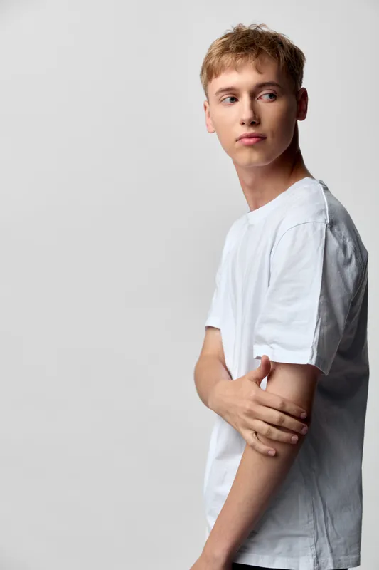Man wearing a white t-shirt in a studio, casual pose with arms crossed and a thoughtful expression against a clean background, lighting creates a simple, focused mood