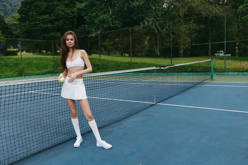 Young female tennis player in a stylish white sports outfit on the court, exuding confidence and determination, surrounded by lush green trees