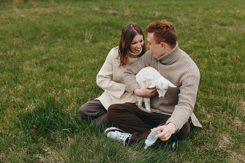 Joyful couple outdoors, sharing a tender moment with a goat kid on a sunlit meadow, cozy sweaters, relaxed posture, gentle breeze, natural colors, intimate connection, rural lifestyle