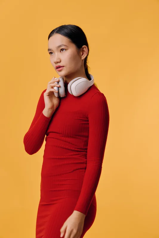 Young woman in red dress with white headphones around neck using portable voice recorder against orange background. Casual portrait of female with neat hairstyle standing and speaking.