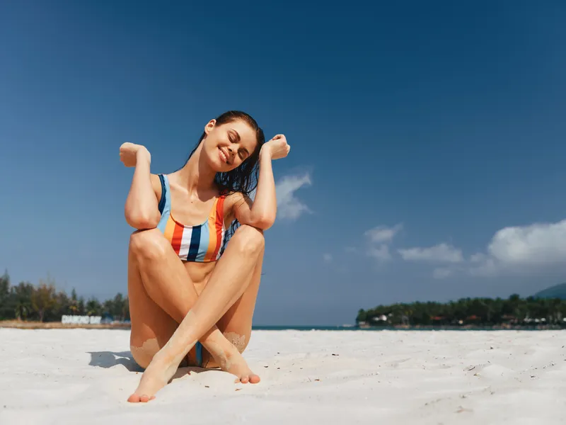 Young woman sits on bright white sand at a sunny beach, wearing a striped swimsuit. She smiles softly with arms raised, radiating relaxed seaside charm.