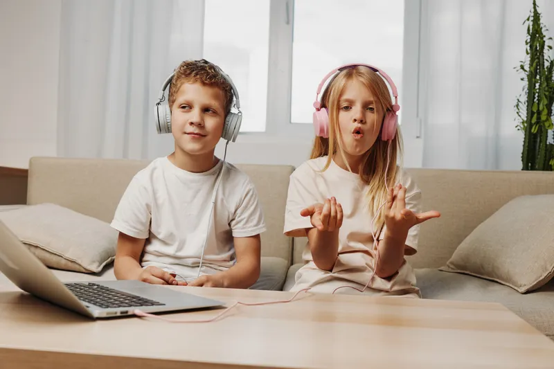 Two children with headphones enjoy music and technology at home, sitting on couch near laptop with bright window behind.