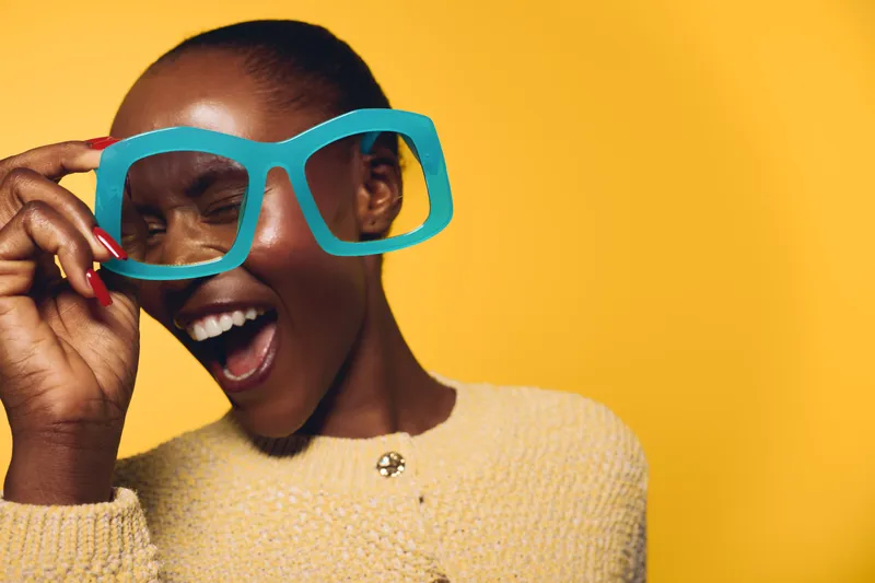 Happy African woman with dark skin wearing oversized blue glasses and beige sweater, laughing joyfully against yellow background. Concept for fun, fashion, and positive emotions.