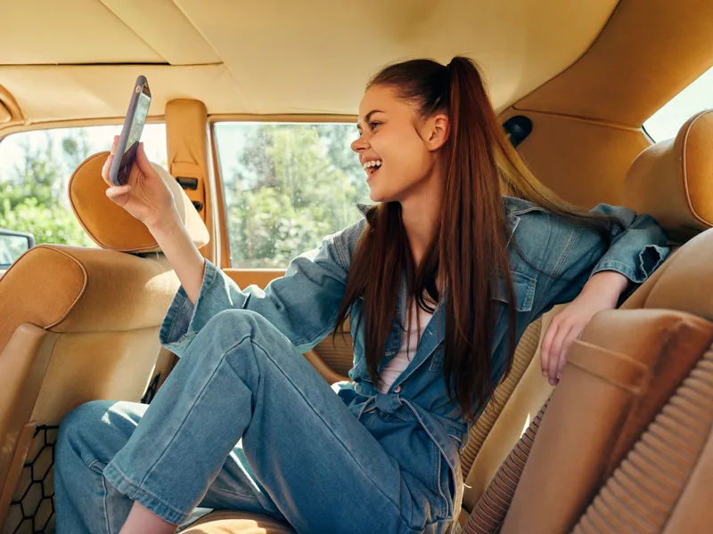 Joyful young woman in denim outfit taking a selfie in a vintage car, showcasing casual style and happiness in a sunny outdoor setting.