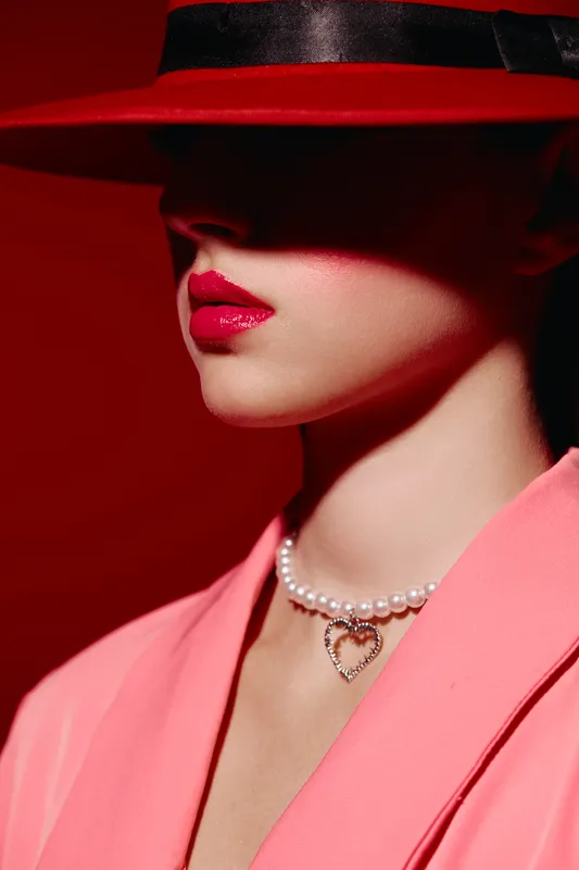 Creative beauty portrait of a woman wearing glossy red lipstick and a pearl necklace with heart pendant. She is partly shadowed by a large red hat, dressed in a pink blazer. Concept for fashion and