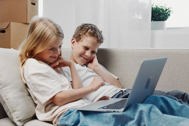 Two children sitting on a couch using a laptop together, smiling and relaxed in a bright living room. Concept of technology, family, childhood, and learning.
