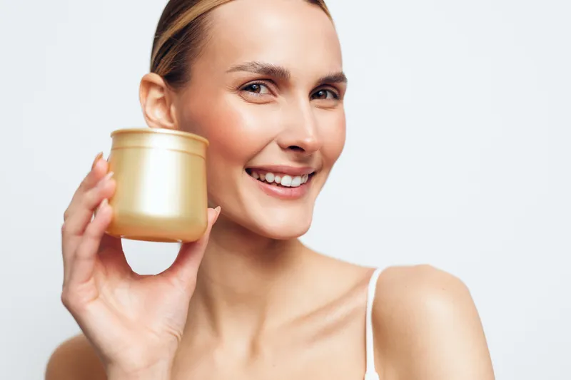 Smiling woman with clear skin holding golden cosmetic jar close to face on white background. Concept of skincare, beauty, wellness, and natural self-care. Soft lighting highlights healthy glow.
