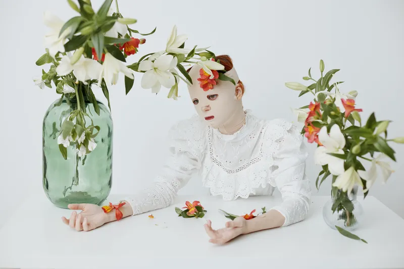 A serene model in a white ruffled dress surrounded by vibrant flowers, expressing tranquility in a minimalist setting, perfect for wellness and beauty concepts