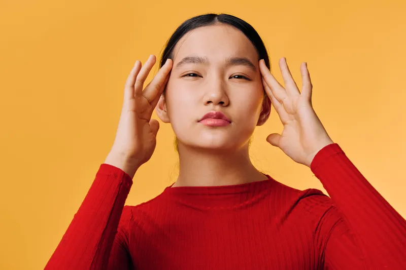 Woman in red sweater with closed eyes touching temples on yellow background, stress or headache concept. Female portrait with natural makeup and minimalist style studio shot.