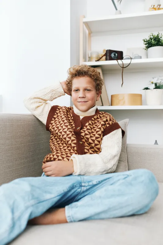 A young boy sits comfortably on a sofa, wearing a patterned knitted sweater, relaxed pose, soft light, and warm tones creating a cozy living room portrait.