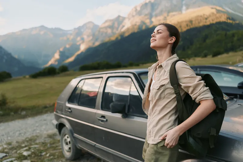 Young woman with backpack stands beside a parked car in a sunlit mountain valley, taking in the view with a relaxed, confident smile as a light breeze brushes her beige shirt