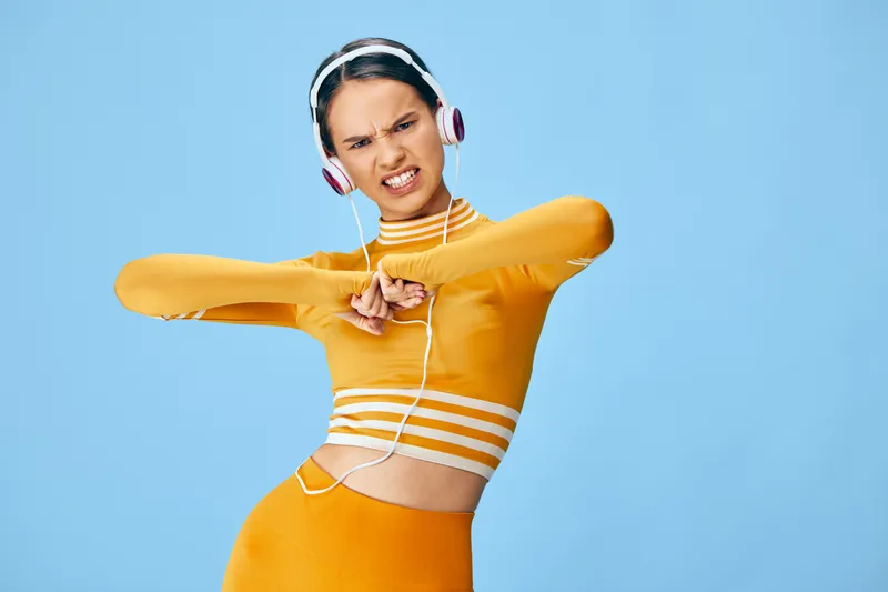 Athletic woman in orange sportswear with headphones, posing mid-dance against a bright blue studio backdrop, capturing energy, movement and motivation in fitness.
