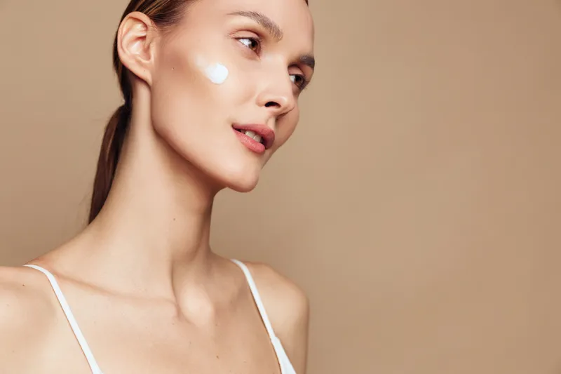 A young woman with clear skin applies white cream on her cheek while looking away. The close-up portrait shows skincare and beauty care concepts with a neutral beige background.