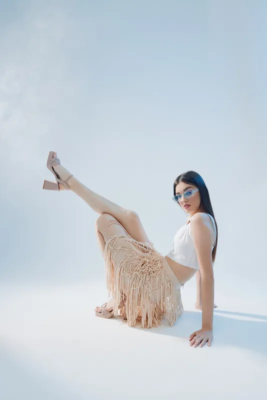 Fashionable woman with long dark hair wearing sunglasses and beige fringe skirt poses on the floor in a bright studio. Concept of beauty, fashion, and style.