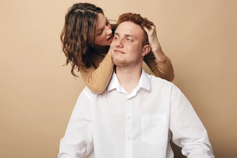 Young couple in casual clothes shows affection, woman hugging man from behind on beige background. Concept of love, relationship, intimacy, and happiness between partners.