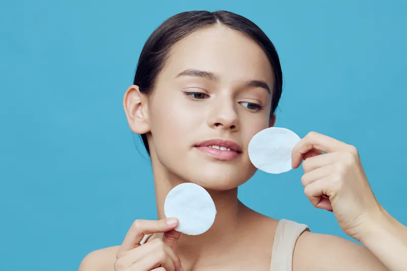 Young woman applying skincare with cotton pads against a blue background, promoting healthy skin and beauty care