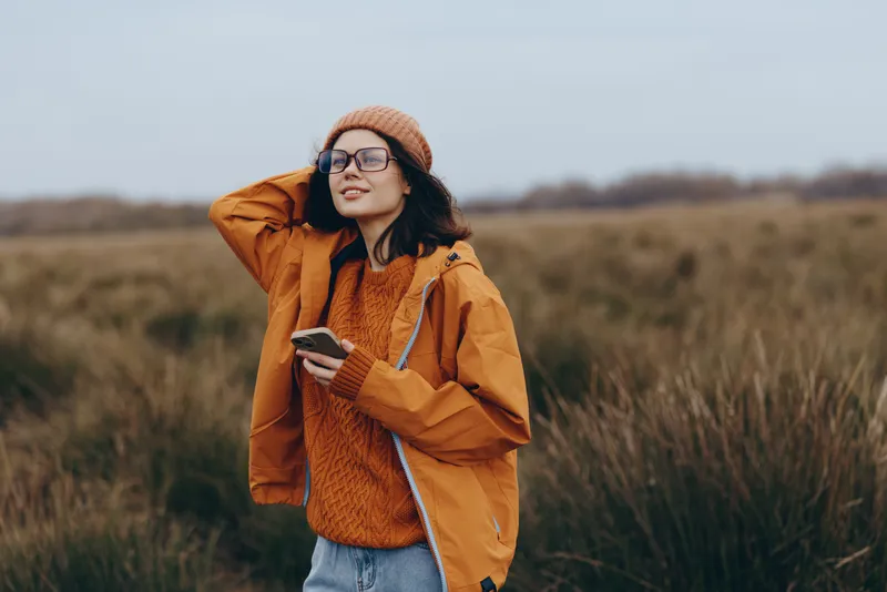 Woman lifestyle outdoors in a grassy field portrait, orange jacket and knit sweater, adjusting hair, holding smartphone, glasses on face, calm mood under cloudy sky
