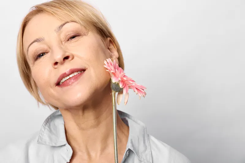 Mature woman smelling a pink flower in spring, closeup portrait, calm smile, nose close to petals suggesting allergies and sensitivity, soft natural light