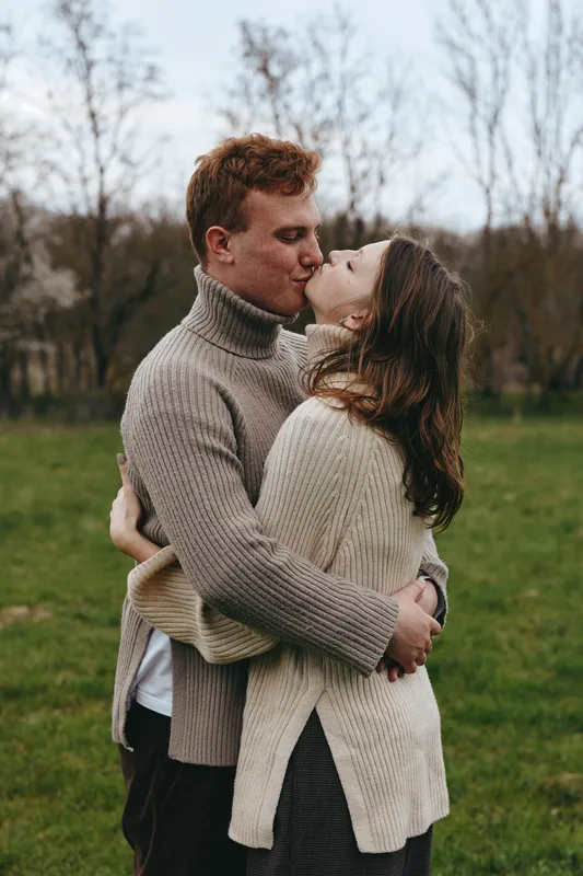 Romantic couple embraces and kisses outdoors in knit sweaters, candid lifestyle shot in a park, warm natural light, affectionate moment, serene field setting, intimate connection.