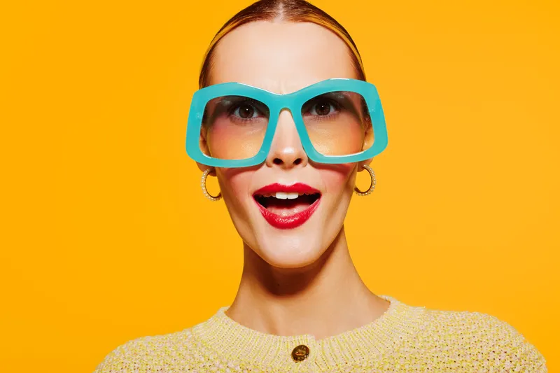 Young woman with bright red lipstick and large blue sunglasses smiles excitedly against vibrant yellow background. Concept of fashion, summer style, and joyful expression.