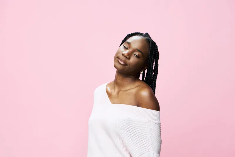Young Black woman with braided hair smiling softly against a pink background, dressed in a cozy white sweater, embodying warmth and confidence