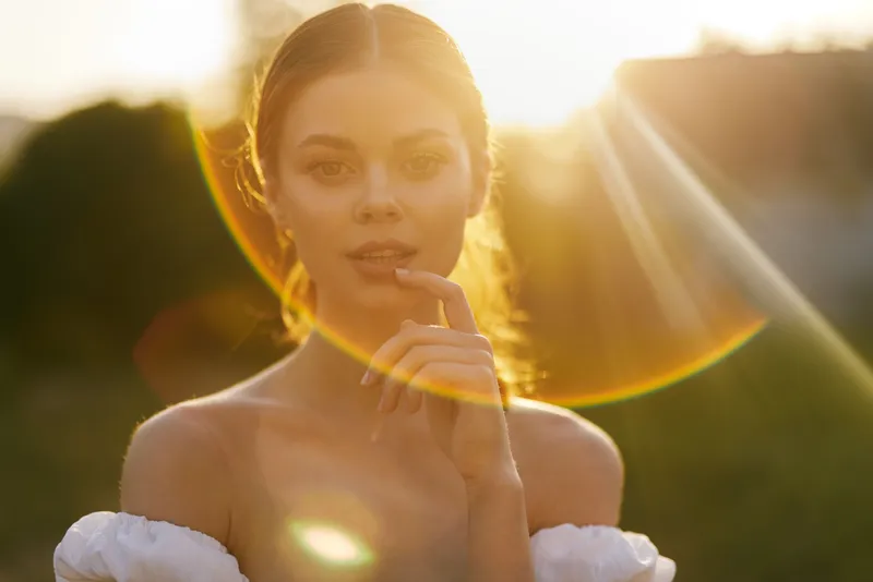 Young woman with natural beauty posing in a sunlit outdoor scene, wearing a white off shoulder dress, evoking feelings of warmth and serenity during golden hour