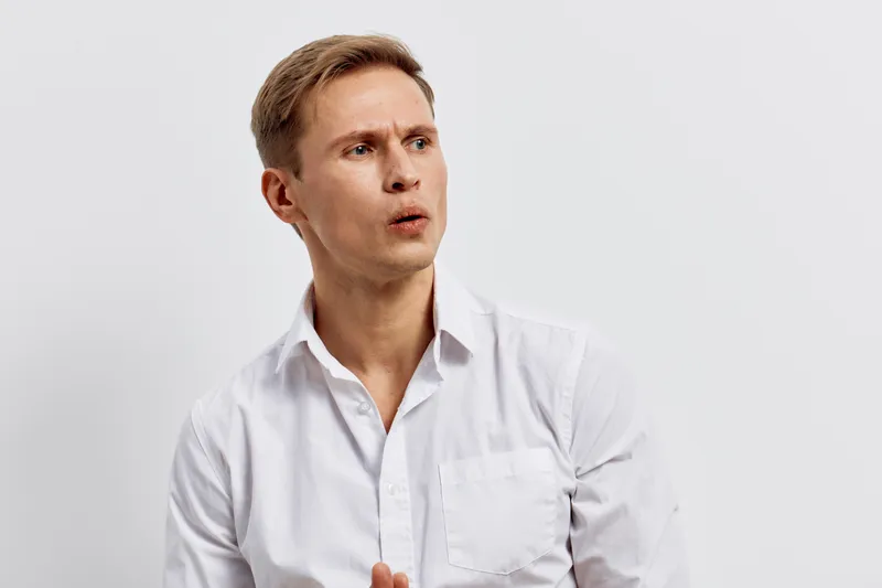 Thoughtful young man with short blond hair wearing a white shirt looking to the side against a plain light gray background studio portrait. People lifestyle concept