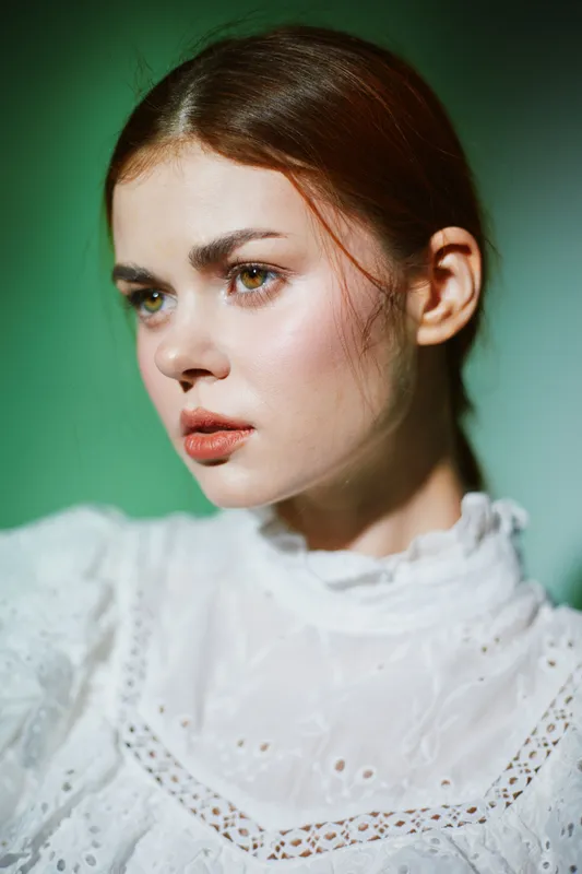 Young woman with a serious expression wearing a white lace blouse against a soft green background, capturing a sense of contemplation and calmness