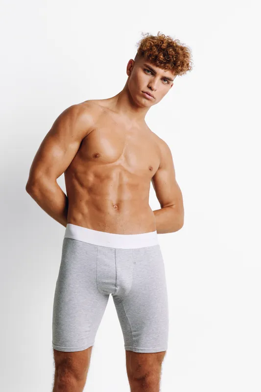 Young muscular man with curly hair posing shirtless in gray shorts against white background. Fitness model showing toned body and confident expression in studio lighting.