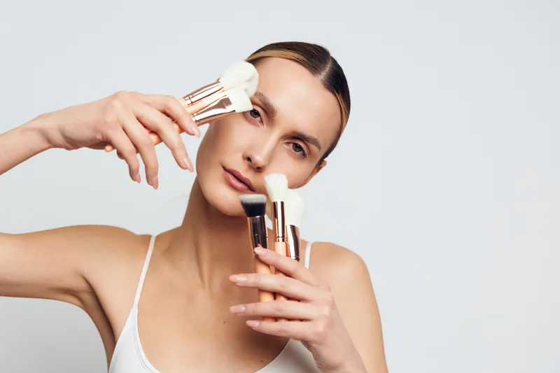 Young woman holds multiple makeup brushes near face applies powder on clean skin with natural expression isolated on light background in beauty concept closeup portrait.