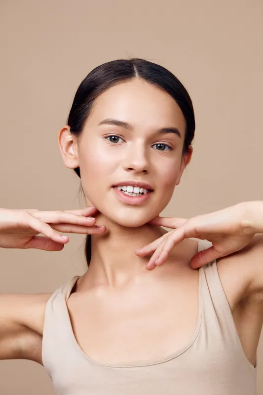 Beautiful young female model with a natural smile showcasing skincare treatment, wearing a nude top against a soft beige background