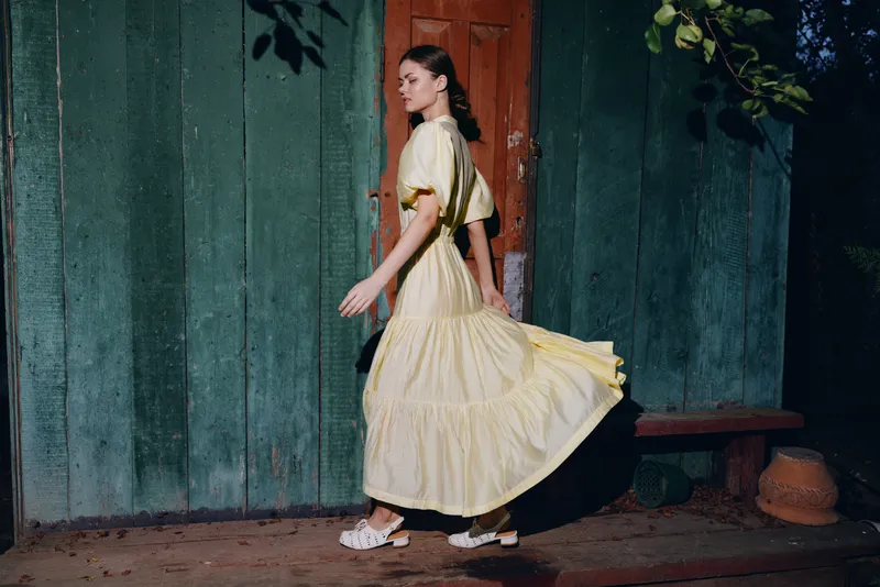 Woman in a flowing yellow dress walks past a weathered green wooden fence at night, creating a cinematic fashion scene with soft lighting and a calm, graceful movement that feels timeless
