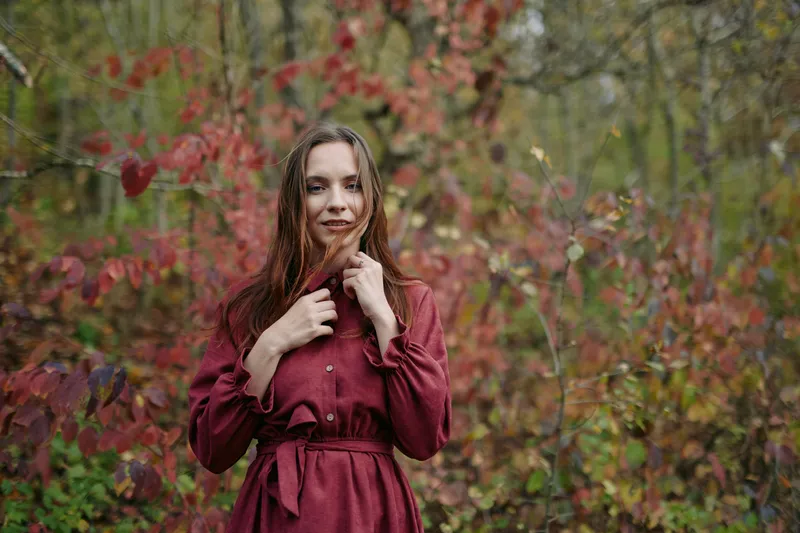 A candid portrait of a woman in a burgundy dress standing in an autumn forest, conveying authenticity and credibility through a natural pose and sincere expression.