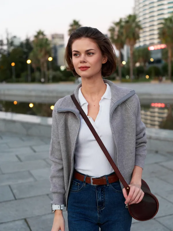 A stylish woman stands outdoors by a waterfront promenade, wearing a light jacket over a white shirt and jeans. She looks thoughtful, confident, and poised in the evening light.