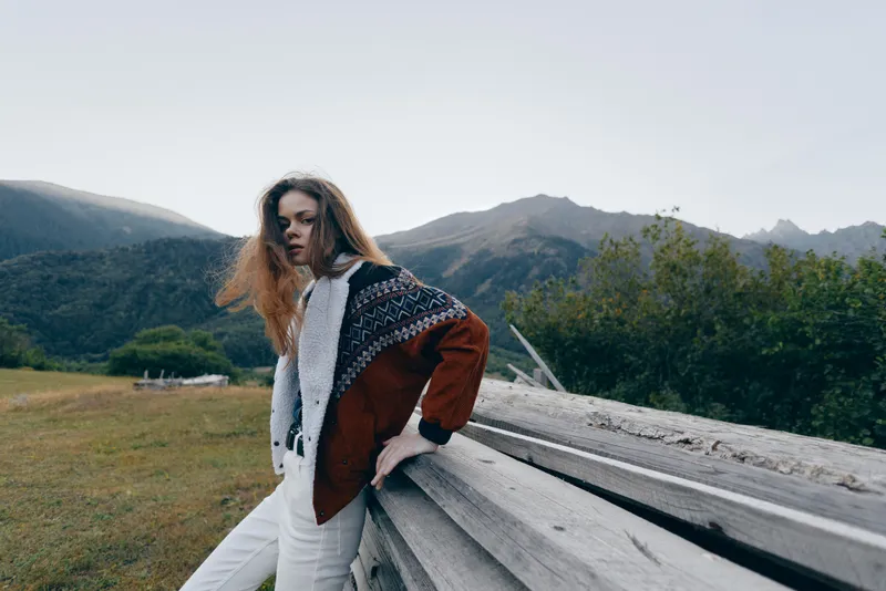 Woman portrait in mountains by wooden fence wearing patterned sweater and jacket, outdoors in nature with fashion attitude, long hair leaning in a serene countryside mood.
