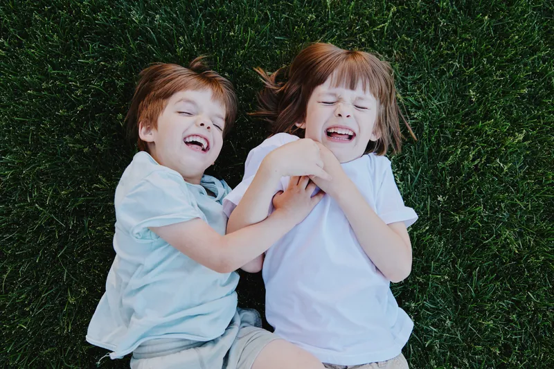 Two joyful children playing on green grass, laughing and smiling together, capturing the essence of childhood happiness in a vibrant outdoor setting.